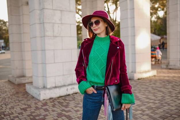 Attraktive Frau im trendigen Herbstoutfit, die auf der Straße spazieren geht und lila Samtjacke, Sonnenbrille und Hut trägt, grüner Strickpullover, Handtasche hält