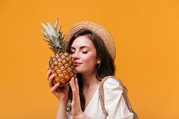 Attraktive Frau im weißen T-Shirt schnüffelt duftende Ananas auf orange Hintergrund.