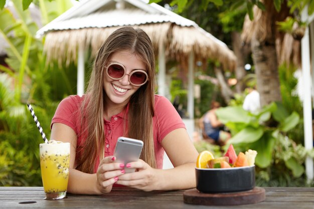 Attraktive Frau in der stilvollen Sonnenbrille, die Smartphone hält und mit glücklichem Blick lächelt, während sie Nachricht von ihrem Freund während des Urlaubs im tropischen Land liest.