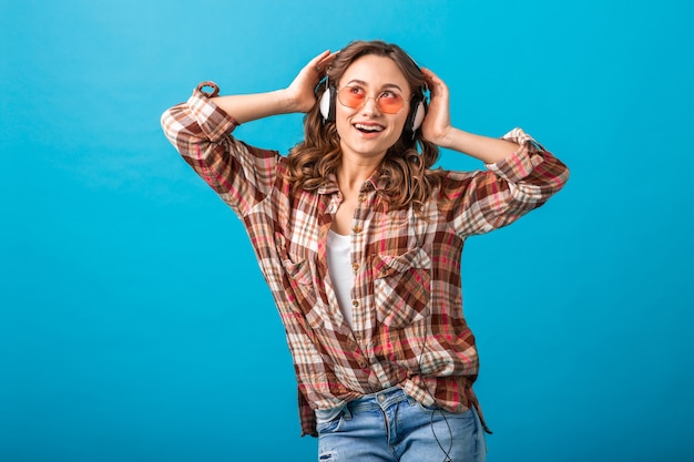 Attraktive Frau lächelnd, die das Hören der Musik in den Kopfhörern im karierten Hemd und in den Jeans lokalisiert auf blauem Studiohintergrund genießt, die rosa Sonnenbrille tragen