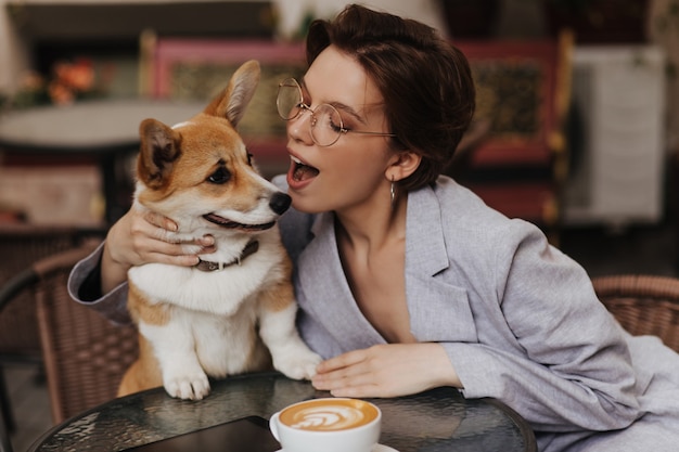 Attraktive Frau mit Brille spielt mit ihrem Hund, während sie sich im Straßencafé ausruht. Charmante Dame in grauer Jacke küsst Corgi