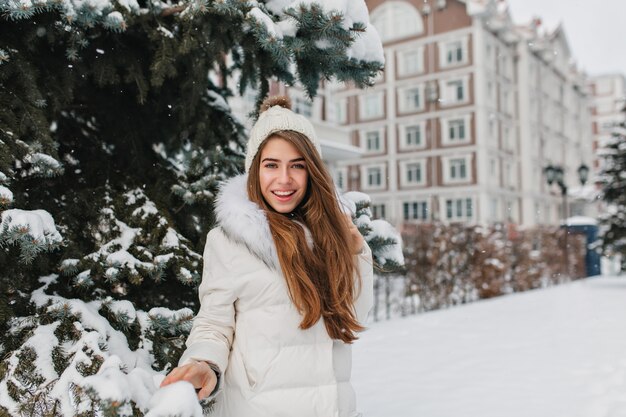 Attraktive Frau mit glattem braunem Haar, das mit selbstbewusstem Lächeln nahe grüner Fichte im Winter aufwirft. Atemberaubende junge Dame trägt weißen Mantel und lustigen Hut, der Spaß mit Schnee hat.