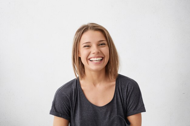 Attraktive Frau mit kurzen blonden Haaren, die sehr froh ist, mit einem breiten Lächeln zu lächeln, das ihre perfekten Zähne zeigt, die Spaß drinnen haben. Freudige aufgeregte fröhliche Frau, die sich freut