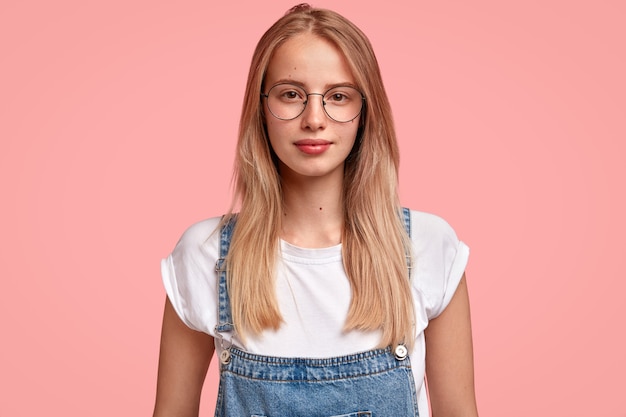 Attraktive Frau mit langen Haaren, angenehmem Aussehen, gekleidet in lässigem T-Shirt mit Jeansoverall und Brille