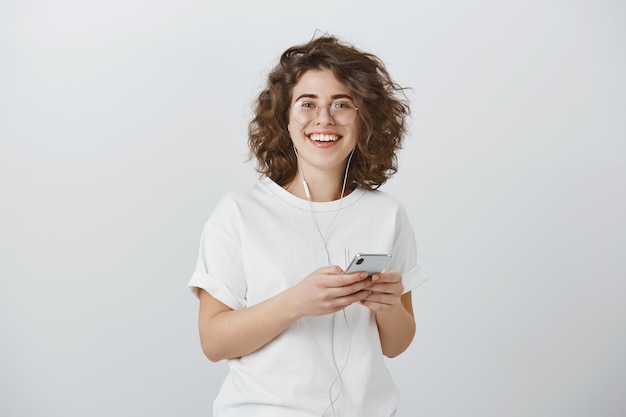 Attraktive freudige junge Frau in der Brille, lächelnd, während sie Smartphone hält und Musik in Kopfhörern hört