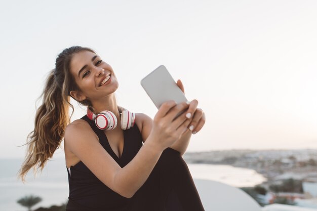 Attraktive freudige junge Frau in der Sportbekleidung lächelnd, am Meer kühlend. Sonnenaufgang am Morgen, modisches Modell, Training, Selfie machen, fröhliche Stimmung