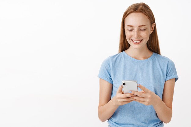 attraktive freundliche und optimistische rothaarige Frau mit Sommersprossen lächelnd und mit Smartphone Blick auf Gerätebildschirm unterhalten