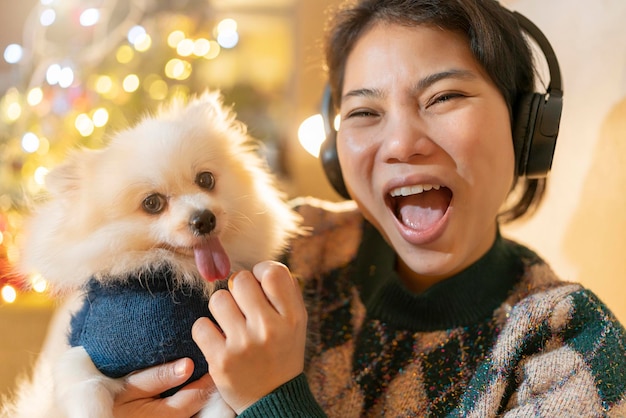 Attraktive, fröhliche asiatische Frauen tragen Wintertuch-Handgriff-Schoßhund mit Pullover-Weihnachtskostüm. Genießen Sie das Sprechen von Foto-Selfie-Smartphone mit Glückslächeln mit Weihnachtsbaum und Lichter-Bokeh