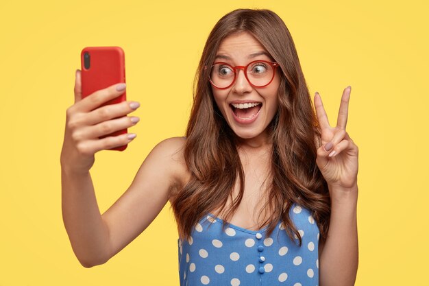 Attraktive fröhliche europäische Frau mit langen Haaren, macht Friedenszeichen, hält Handy, posiert für Selfie, gekleidet in gepunktetes blaues Kleid, isoliert über gelbe Wand macht Blog über den Alltag