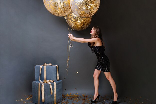 Attraktive fröhliche junge Frau im schwarzen Luxuskleid, die Spaß mit großen Luftballons voll mit goldenen Lametta hat. Alles Gute zum Geburtstag, Geschenke, Lächeln, Positivität ausdrücken.