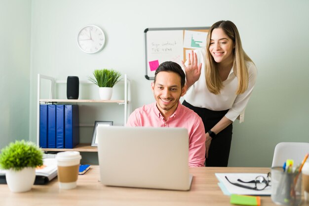 Attraktive Geschäftsleute, die ein Online-Arbeitstreffen mit einem Kunden im Büro haben. Lächelnde kaukasische und hispanische Kollegen bei einem Videoanruf an einem Laptop