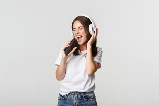 Attraktive glückliche Frau, die Karaoke-App spielt, in Smartphone singt, drahtlose Kopfhörer trägt.