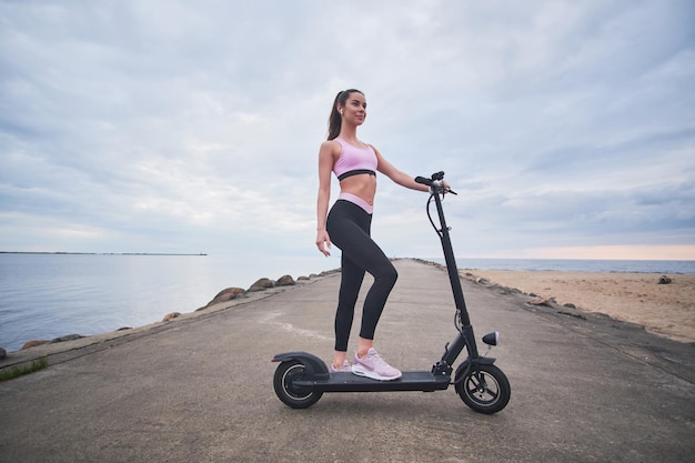 Attraktive glückliche Frau genießt das morgendliche Reiten mit ihrem neuen Elektroroller am Meer.