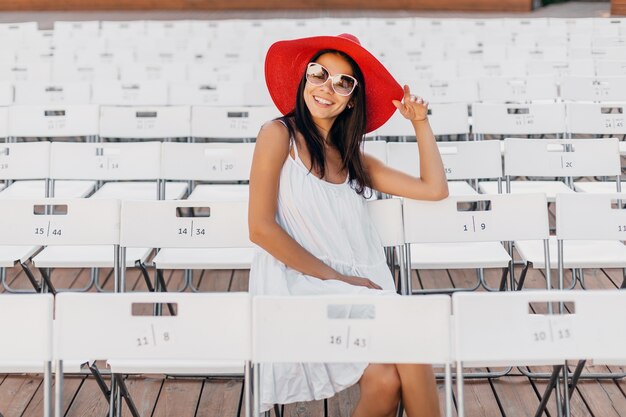 Attraktive glückliche lächelnde Frau gekleidet in weißem Kleid, rotem Hut, Sonnenbrille, die im Sommer Freilufttheater auf Stuhl allein, Frühlingsstraßenart-Modetrend sitzt