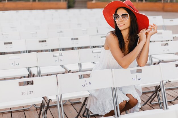 Attraktive glückliche lächelnde Frau gekleidet in weißem Kleid, rotem Hut, Sonnenbrille, die im Sommer Freilufttheater auf Stuhl allein, Frühlingsstraßenart-Modetrend sitzt