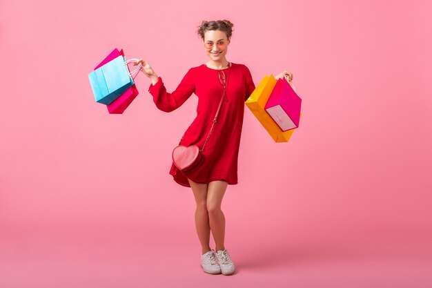 Attraktive glückliche lächelnde stilvolle Frau shopaholic im roten trendigen Kleid, das bunte Einkaufstaschen auf rosa Wand lokalisiert, Verkauf aufgeregt, Frühlingssommermodetrend hält