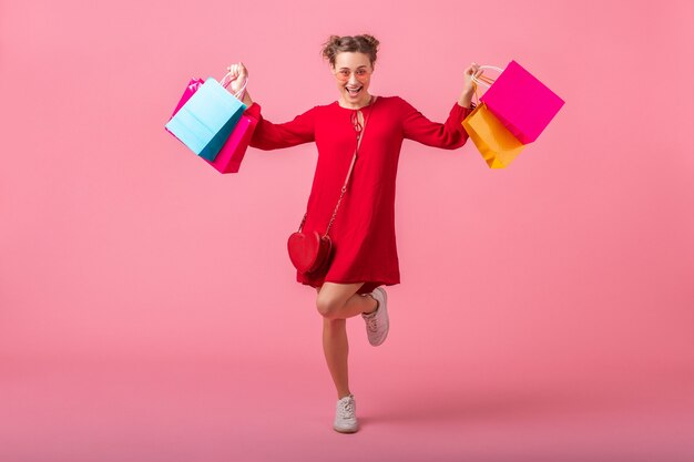 Attraktive glückliche lächelnde stilvolle Frau shopaholic im roten trendigen Kleid, das bunte Einkaufstaschen auf rosa Wand lokalisiert, Verkauf aufgeregt, Frühlingssommermodetrend hält