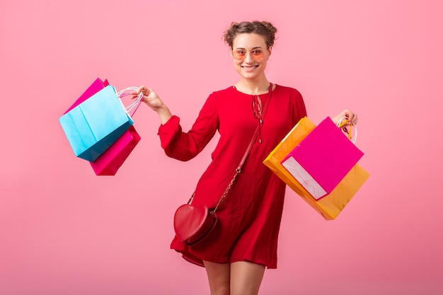 Attraktive glückliche lustige emotion stilvolle frau shopaholic im roten trendigen kleid, das bunte einkaufstaschen auf rosa wand lokalisiert, verkauf aufgeregt, frühlingssommer-modetrend hält