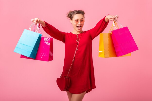 Attraktive glückliche lustige Emotion stilvolle Frau shopaholic im roten trendigen Kleid, das bunte Einkaufstaschen auf rosa Wand lokalisiert, Verkauf aufgeregt, Frühlingssommer-Modetrend hält