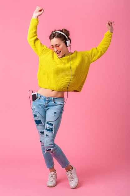 Attraktive glückliche lustige Frau, die Musik hört, die in den Kopfhörern gekleidet ist, gekleidet in buntem Hipster-Stil-Outfit lokalisiert auf rosa Studiohintergrund