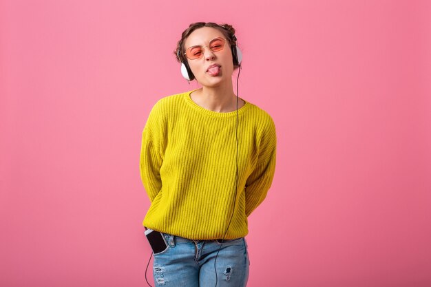 Attraktive glückliche lustige Frau, die Musik in Kopfhörern gehört, gekleidet in buntem Hipster-Stil-Outfit lokalisiert auf rosa Wand, tragenden gelben Pullover und Sonnenbrille, Spaß habend