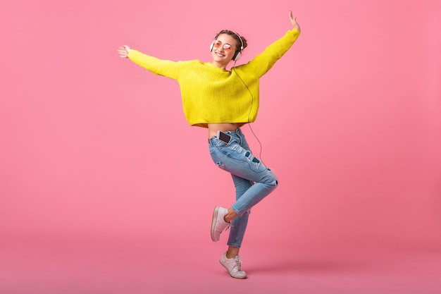 Attraktive glückliche lustige Frau, die springend hört Musik in Kopfhörern gekleidet in buntem Hipster-Stil-Outfit lokalisiert auf rosa Wand, tragenden gelben Pullover und Sonnenbrille, Spaß habend