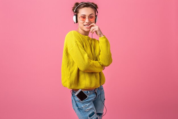 Attraktive glückliche lustige lustige flirty Frau, die Musik in Kopfhörern gekleidet im bunten Hipster-Stil-Outfit lokalisiert auf rosa Wand hört, gelben Pullover und Sonnenbrille tragend, Spaß hat