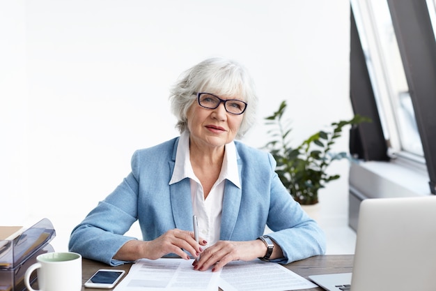 Attraktive grauhaarige Senior-Geschäftsfrau in modischem Anzug und Brille, die in ihrem Büro arbeitet, mit offenem Laptop und Papieren am Schreibtisch sitzt, Finanzdokumente ausfüllt und einen ernsthaften Blick hat