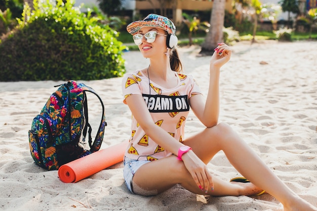 Attraktive Hipster-Frau, die am Strand sitzt und Musik auf Kopfhörern in stilvollem buntem Outfit auf tropischen Sommerferien hört, die Zubehörkappen-Sonnenbrille tragen und lächelnd mit Rucksack reisen