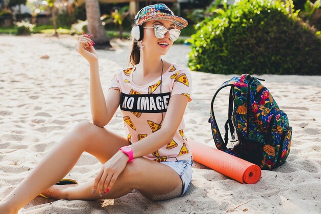 Attraktive Hipster-Frau, die am Strand sitzt und Musik auf Kopfhörern in stilvollem buntem Outfit auf tropischen Sommerferien hört, die Zubehörkappen-Sonnenbrille tragen und lächelnd mit Rucksack reisen