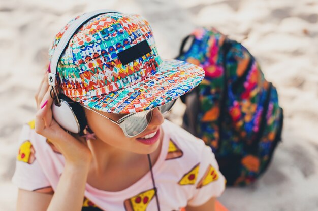 Attraktive Hipster-Frau, die auf Strand geht, die Musik auf Kopfhörern in stilvollem buntem Outfit auf tropischen Sommerferien hört, die Zubehörkappen-Sonnenbrille tragen, lächelnd reisen mit Rucksack