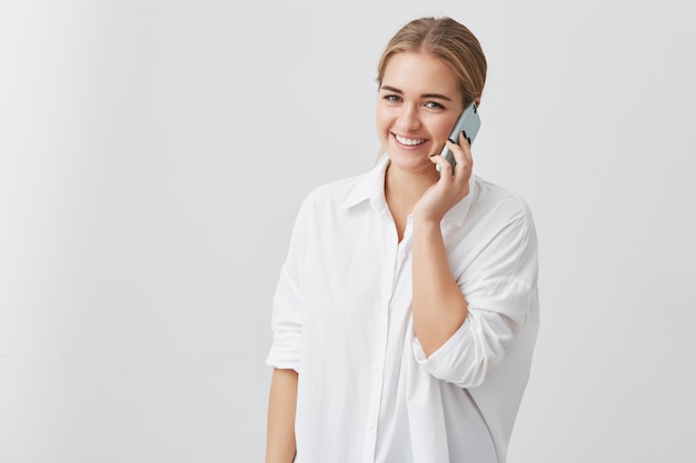 Attraktive hübsche blonde Frau, die weißes Hemd trägt, das anmutiges und glückliches Aussehen beim Sprechen über Smartphone mit ihrem Geliebten hat. Menschen- und Technologiekonzept