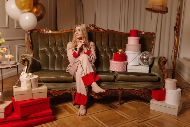 Attraktive junge blonde kaukasische Dame im Schlafanzug sitzt auf dem Sofa im dekorierten festlichen Raum Weihnachtsvorbereitungskonzept