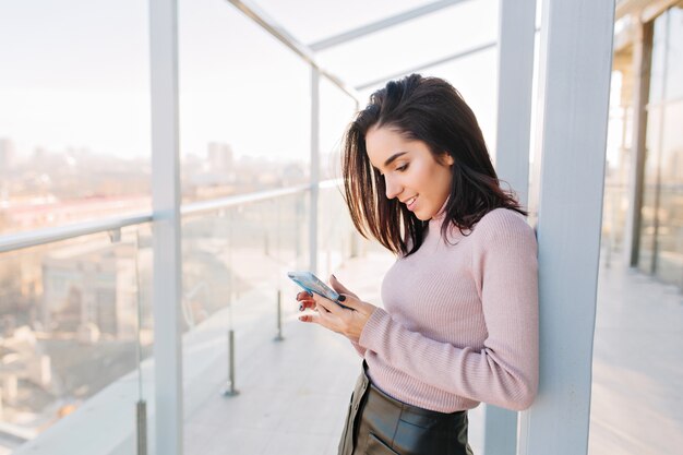 Attraktive junge brünette Frau, die am Telefon auf Terrasse auf Großstadtansicht tippt. Sonniger Morgen, entspannte Zeit, fröhliche Gefühle, charmante, elegante Aussicht.