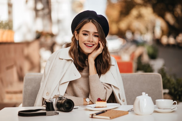 Attraktive junge Dame mit brünetter, gewellter Frisur, Baskenmütze, beigem Trenchcoat, die auf der Café-Terrasse gegen die sonnige Herbststadtmauer zu Mittag isst