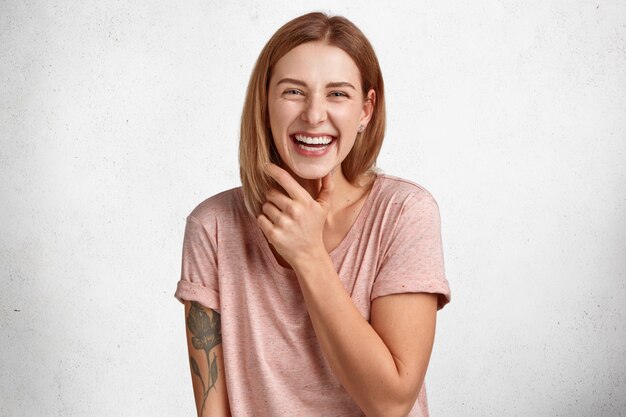 Attraktive junge europäische Frau, die sehr froh ist, perfekte Zähne zeigt, drinnen Spaß hat, sich nach dem Vorschlag zur Fröhlichkeit freut, Hand tätowiert hat, isoliert über weißer Betonwand. Glückskonzept
