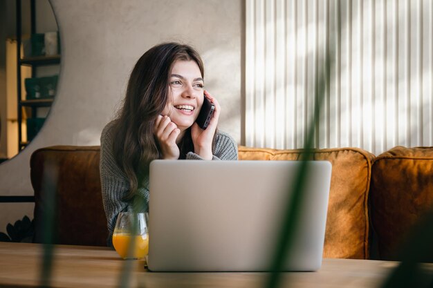 Attraktive junge Frau, die am Telefon spricht und an einem Laptop arbeitet