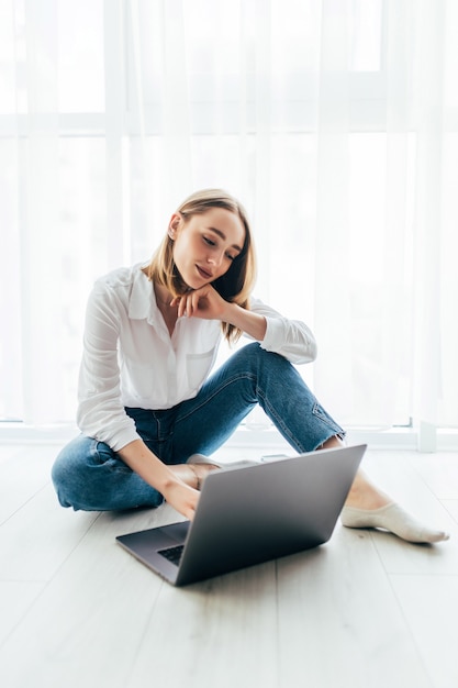 Attraktive junge Frau, die auf ihrem Laptop auf dem Boden sitzt surft