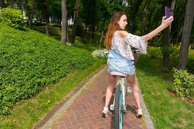 Attraktive junge frau, die ein selfie mit ihrem smartphone macht, während sie auf ihrem fahrrad in einem park sitzt