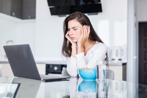 Attraktive junge Frau, die Laptop beim Frühstück benutzt und in der Küche sitzt.