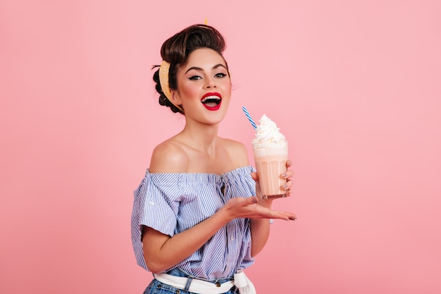 Attraktive junge Frau, die Milchshake genießt. Pinup-Mädchen in gestreifter Bluse, die auf rosa Hintergrund aufwirft.