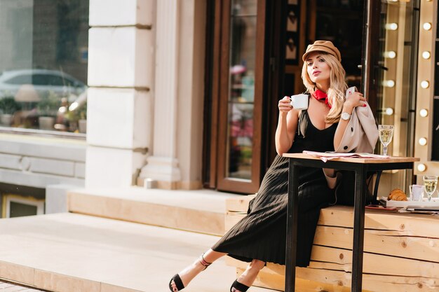 Attraktive junge Frau, die nach der Arbeit im Lieblingscafé ruht und Kaffeegeschmack genießt. Außenporträt des blonden Mädchens im stilvollen Outfit, das am Wochenende entspannt.