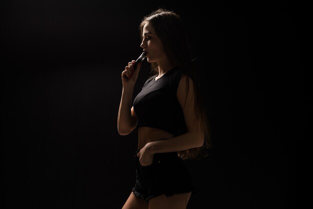 Attraktive junge Frau, die Rauch verdampft und bläst, lokalisiert auf schwarzer Wand