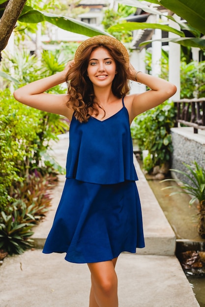 Attraktive junge Frau im blauen Kleid und im Strohhut, die im tropischen Spa-Villenhotel im Urlaub im Sommerart-Outfit, sexy gehen