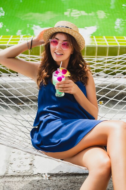 Attraktive junge Frau im blauen Kleid und im Strohhut, die rosa Sonnenbrille trinkenden Alkoholcocktail im Urlaub sitzen in der Hängematte im Sommerart-Outfit, glücklich in der Party-Stimmung lächelnd