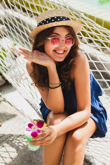 Attraktive junge Frau im blauen Kleid und im Strohhut, die rosa Sonnenbrille trinkenden Alkoholcocktail im Urlaub sitzen in der Hängematte im Sommerart-Outfit, glücklich in der Party-Stimmung lächelnd