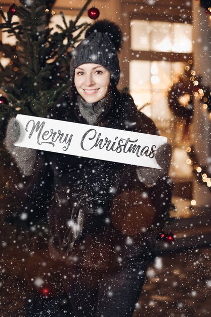 Attraktive junge Frau in warmen Kleidern steht unter dem Schnee und wünscht allen frohe Weihnachten
