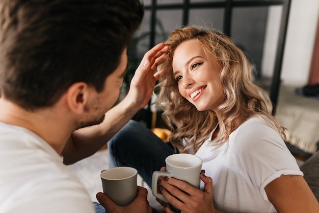 Attraktive junge Frau mit blonden Haaren, die ihren Freund ansieht und lächelt, während er ihre Haare repariert. Glückliches Paar in der Liebe, die Zeit zu Hause zusammen verbringt.