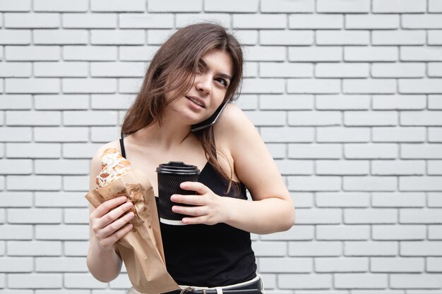 Attraktive junge Frau mit einem Croissant und Kaffee vor einer weißen Backsteinmauer