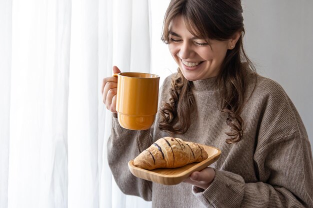 Attraktive junge Frau mit einer Tasse Heißgetränk und einem Croissant am Fenster